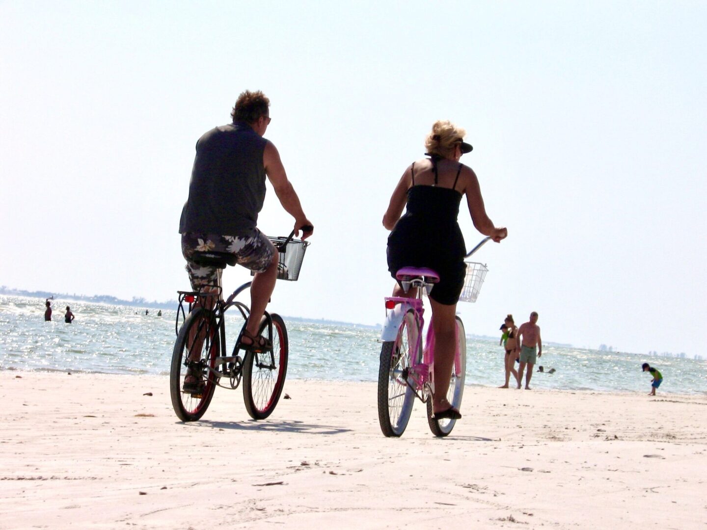 Two people riding bikes on the beach near water.